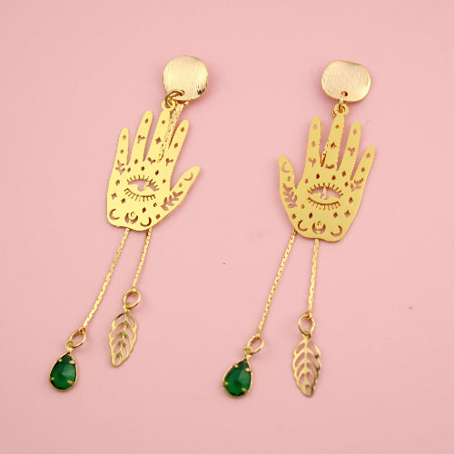 Gold hand-shaped earrings with green gemstones on a pink background
