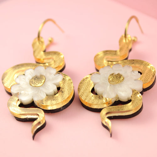 Gold earrings with white floral designs on a pink background