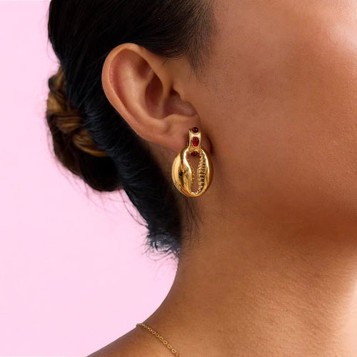 Gold earring with red gemstone worn by a person against a pink background