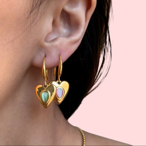 Gold heart-shaped earrings with colorful stones worn by a person against a pink background