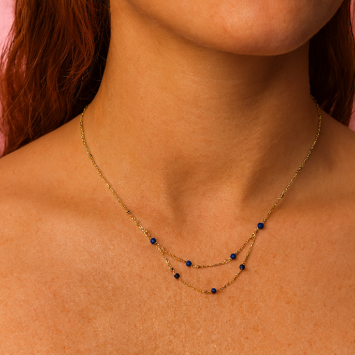Close-up of a person wearing a gold necklace with blue beads on a pink background