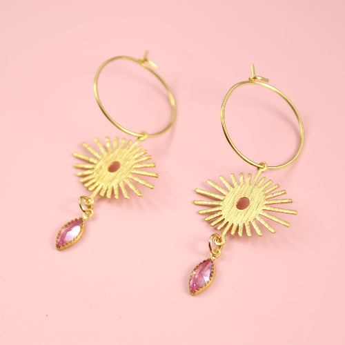 Gold earrings with sunburst designs and pink gemstones on a pink background