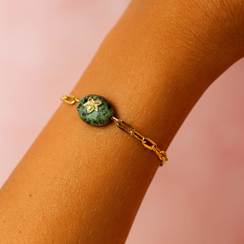 Gold bracelet with a green gemstone on a wrist against a pink background